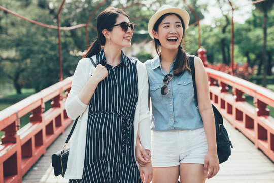 two asian girl friends on walking on beautiful wooden bridge in japanese traditional park. red small arched bridge over stream. young women on walk way crossing pond outdoor on sunny day talk smiling