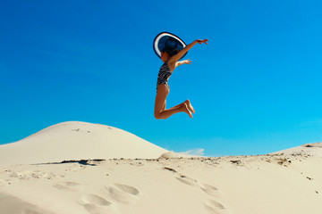 Happy young girl jumping in a desert.  People, happiness, travel concept. Teenage girl wearing a big black hat jumping on a beach. Attractive girl jumping on the beach over blue sky background.