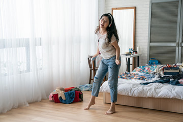 Beautiful woman packing suitcase in cozy bedroom. full length happy young asian female standing on wooden floor hold mobile phone with headphones dancing in modern house. cheerful funny girl pose - obrazy, fototapety, plakaty