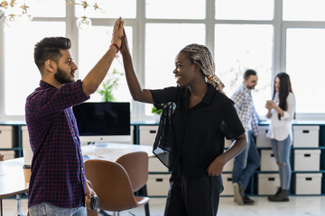Celebrating success. Cheerful young indian man and african woman giving high-five while having coffee break in creative office