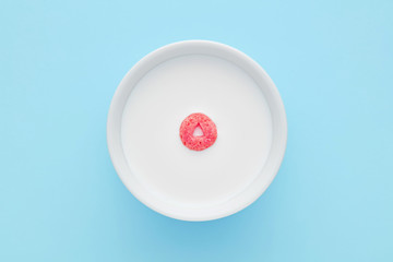 Single cereal ring floating on a white bowl full of milk on blue background. Close up top down view.