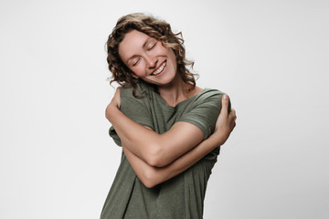 Young curly woman hugging herself, looks happy, expresses natural positive emotions