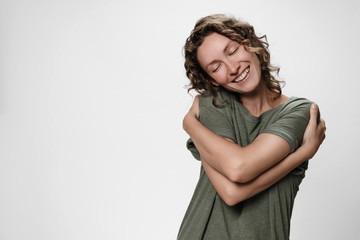 Young curly woman hugging herself, looks happy, expresses natural positive emotions