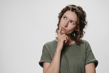 Puzzled curly woman holds chin, looks thoughtfully upwards, being deep in thoughts