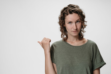Caucasian curly woman with friendly expression, points aside with thumb, shows free copy