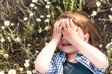 cute little boy closes his eyes with his hands lying in a daisy field
