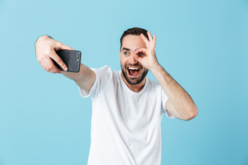 Photo of unshaved brunette man wearing basic t-shirt laughing and taking selfie on smartphone