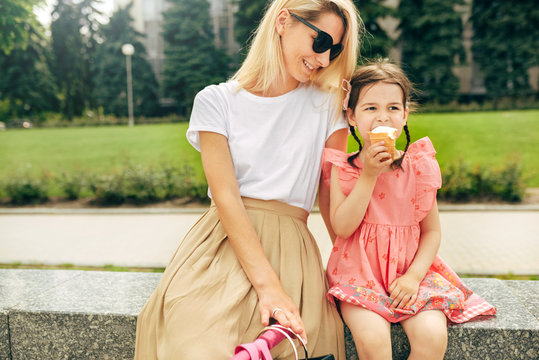 Horizontal Outdoors Image Of Happy Cute Little Girl Sitting With Mother On The City Street And Eating Ice-cream. Fun Girl Kid And Mom Having Fun And Playing Outside. Motherhood And Childhood Concept