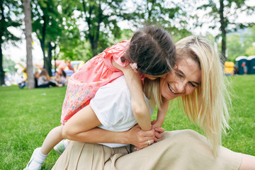 Cheerful child embracing mother in the park. Pretty daughter piggybacks her young attractive mother sitting on green grass outside. Love emotion. Happy Mothers Day. Motherhood and childhood concept
