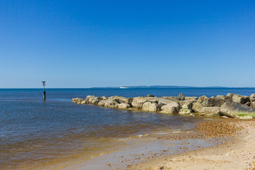 A view of a sandy beach with a stony groyne (breakwater), calm crystal sea and cormorant spreading his wings on the weathercock under a majestic blue sky
