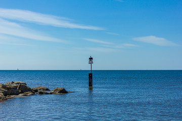 Fototapeta premium A view of a weathercock in the end of a stony groyne (breakwater) with a cormorant spreading his wing and crystal blue water under a majestic blue sky and some white clouds