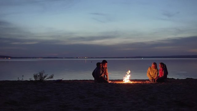 Family Of Travelers With Kids Rest Around Night Campfire By Sea