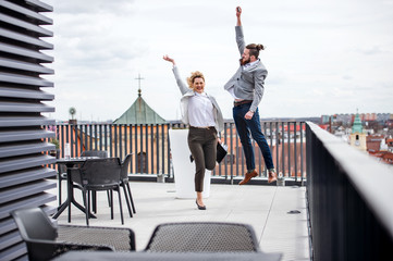 Two young business people jumping on terrace outside office, expressing excitement.