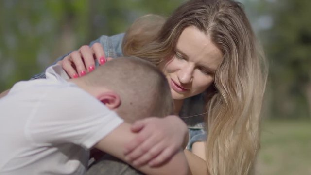 The little boy sitting on the grass hiding his face, a young beautiful woman caressing his head, trying to cheer him up. A mother comforts her son. A loving family, support in a difficult situation