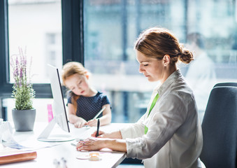 A businesswoman with small daughter sitting in an office, working.