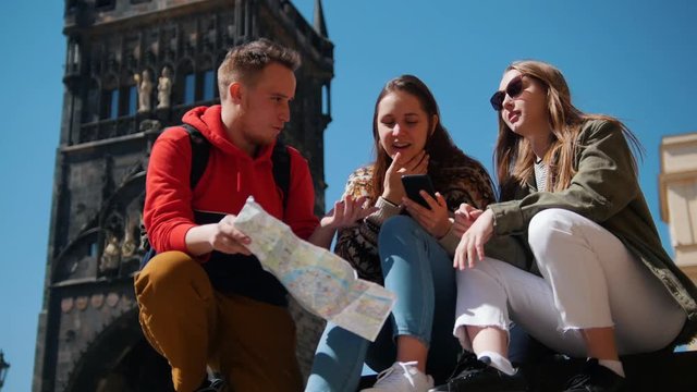 Young friends travelers sitting on a background of Bridge Tower of Charles Bridge and searching for a way to go in their phone navigator and on the map