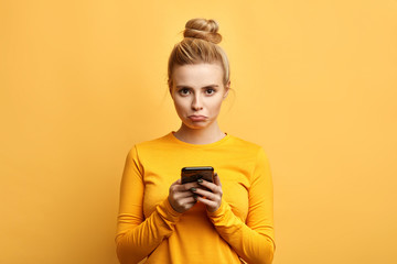 Fototapeta angry sad woman with hairbun annoyed by something while using phone, girl has received bad sms, text message isolated yellow background. emotion, reaction, feeling obraz