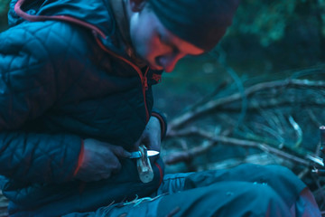 Young man in cap cutting wood near campfire.