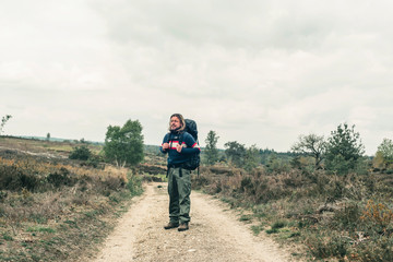 Obraz premium Man with long blonde hair and red backpack on dirt road in hilly landscape under cloudy sky.