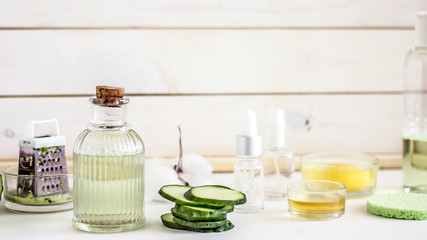 Homemade cucumber lotion in a transparent bottle with cork stopper stands on a wooden white table, next to slices of fresh cucumber. In the background are ingredients for making homemade cosmetics. 