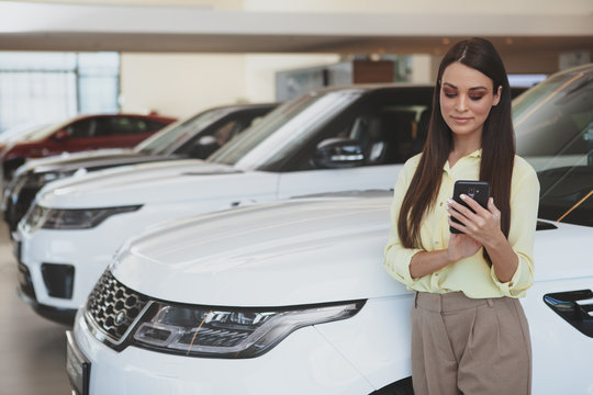 Attractive Female Cusotmer Using Smart Phone While Choosing New Car To Buy At Dealership Showroom. Beautiful Businesswoman Shopping For New Auto, Copy Space