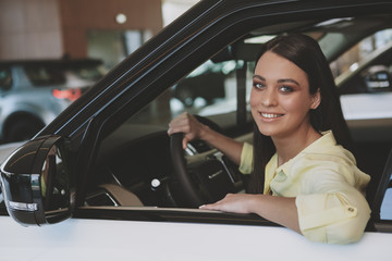 Obraz na płótnie Canvas Happy attractive young woman smiling to the camera, sitting in a new car holding steering wheel. Cheerful businesswoman examining new automobile for sale at the dealership