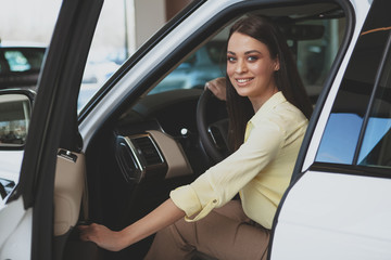 Fototapeta na wymiar Excited young woman smiling to the camera, getting in a new automobile at the dealership. CHeerful beautiful woman sitting in a new car, choosing auto to buy at cars salon