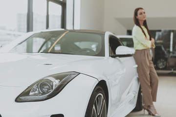 Selective focus on car lights, elegant woman leaning on the automobile, looking away dreamily. Attractive woman buying new car at the dealership, copy space. Travelling, tourism concept