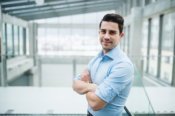 A portrait of young businessman standing indoors in an office, arms crossed.
