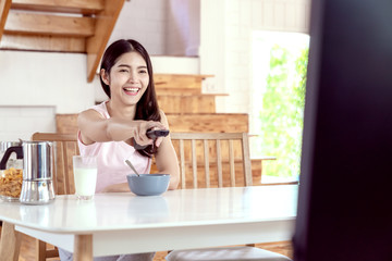 Candid of young happy attractive asian girl enjoy weekend activity by watching TV program at home. Young asian female student sitting on desk table holding remote on hand and pointing toward to TV.