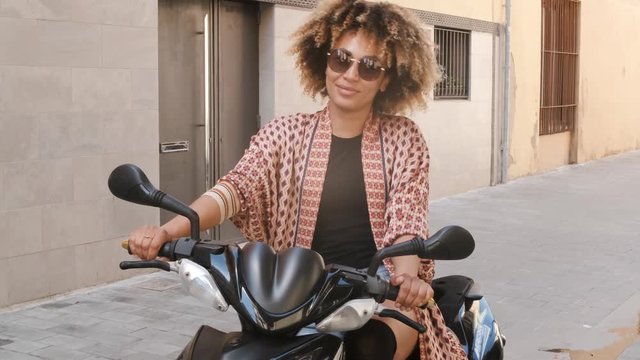 Trendy ethnic woman sitting on scooter on street