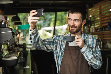 Portrait of brunette barista man taking selfie photo while working in street cafe or coffeehouse outdoor