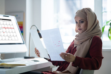 Dark-eyed teacher wearing hijab sitting near computer