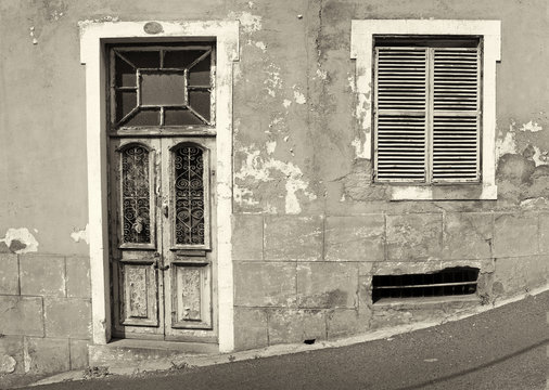 the front of an old abandoned house with shuttered windows and locked wooden door with flaking peeling paint on a sloping street monochrome image