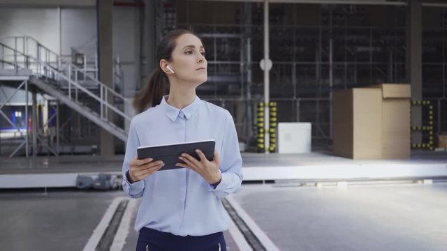 Female manager talking on wireless headphones while using tablet and supervising work in factory