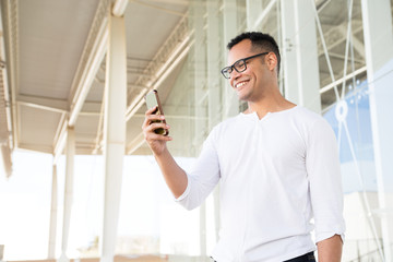 Bottom view of smiling young mixed-race man in spectacles and white T-shirt standing at office building, holding phone in hands, looking aside. Lifestyle concept
