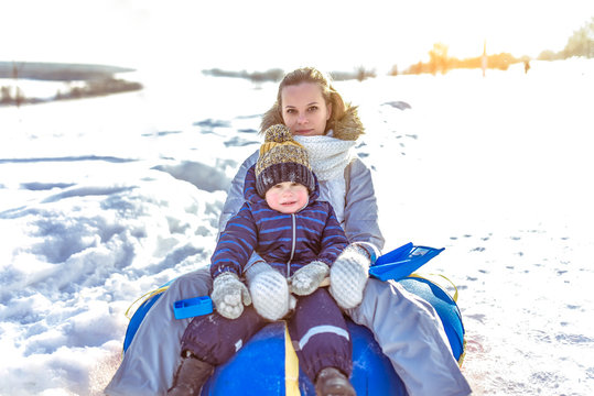 Mom woman with child boy 3 years old, sitting on inflatable tubing, rolling down slide resting winter in fresh air weekend vacation resort, background snow drifts. In his hand a toy snow mat.