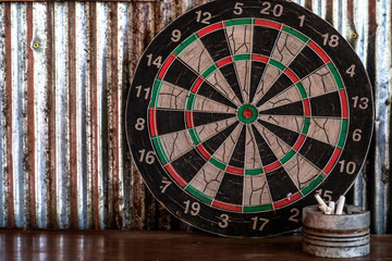 A dart board leaning against silver galvanized iron wall with cigarettes in a zinc tin ashtray in front.