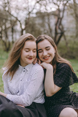Fototapeta premium Portrait of two beautiful young sisters sitting in the green spring park, hugging in the field. Having fun together, positive emotions, bright colors. Copy space. Happy girlfriends at sunset
