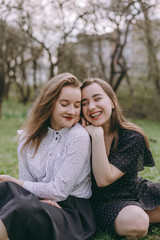 Portrait of two beautiful young sisters sitting in the green spring park, hugging in the field. Having fun together, positive emotions, bright colors. Copy space. Happy girlfriends at sunset