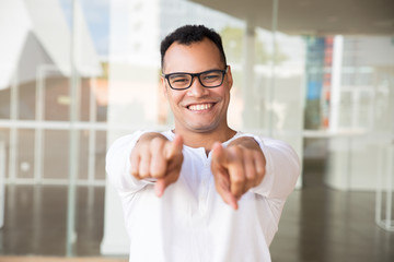 Fototapeta na wymiar Medium shot of smiling young mixed-race man in spectacles and white T-shirt standing at office building, looking straight at camera, pointing at the camera with both hands. Lifestyle concept