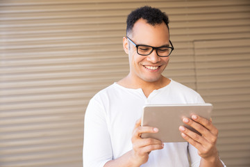 Medium shot of smiling young mixed-race man in spectacles and white T-shirt standing at office building, working on tablet. Front view. Lifestyle concept