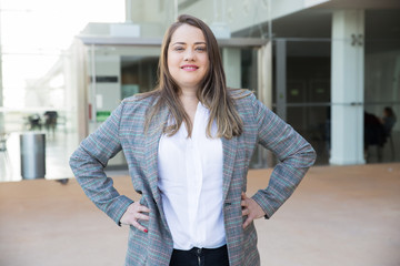 Smiling business woman keeping hands on hips outdoors. Lady looking at camera and standing with building in background. Business lady portrait concept. Front view.