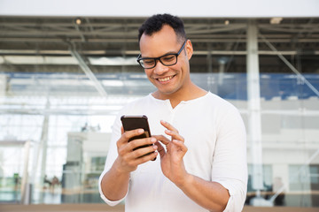 Smiling young man texting on smartphone outdoors. Handsome guy standing with building glass wall in background. Communication concept. Front view.