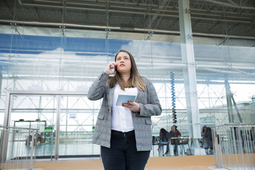 Focused female employee discussing project on phone in office lobby. Young woman in formal jacket standing against glass wall, holding tablet and talking on cellphone. Communication concept