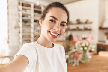 Image of smiling charming woman taking selfie photo and winking in cozy kitchen