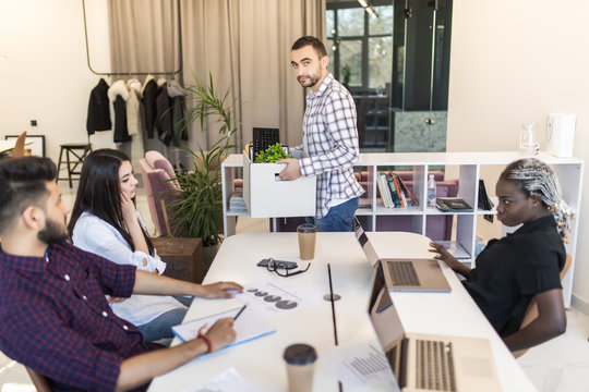Young sad employee with personal belongings standing in the office and say goodbay to the team while change his workplace.