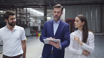 Manager walking in factory with two engineers explaining and showing on tablet with forklift driving in background