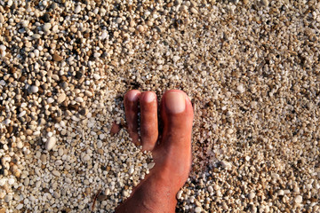 Top view of man standing bare feet on beach. Texture of bottom, leg and foot of man drowning with sweeping colorful stone below on rocky stones beach in sea with waves and foam, water ripples of sea.