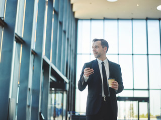 Dreamy middle aged businessman in suit looking through window while walking along lobby in office building holding smartphone and coffee cup in his hands